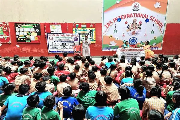 स्कूल परिसर में मंच के सामने बैठे विद्यार्थियों को संबोधित करती महिला और पास लगी बाल सुरक्षा जागरूकता की तख्ती