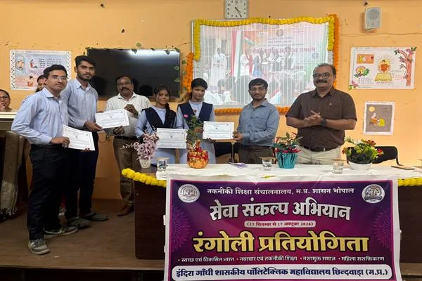 मंच पर प्रमाणपत्र लिए खड़े छात्र-छात्राएं और शिक्षक, सामने रंगोली प्रतियोगिता का बैनर तथा मेज पर सजी रंगोलियां