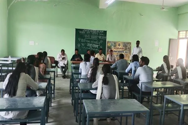 कक्षा में डेस्क पर बैठे छात्र-छात्राओं के सामने मंच पर मौजूद महाविद्यालय के शिक्षक और वक्ता