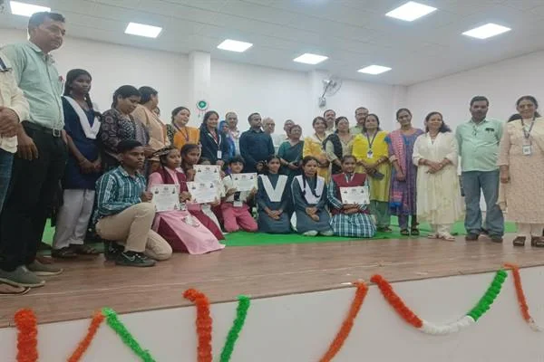 एक हॉल के मंच पर छात्र-छात्राएं प्रमाण-पत्र हाथ में लिए बैठे हैं, पीछे शिक्षक और अधिकारी समूह में खड़े हैं; मंच के आगे तिरंगे रंग की फूलमाला लगी है।