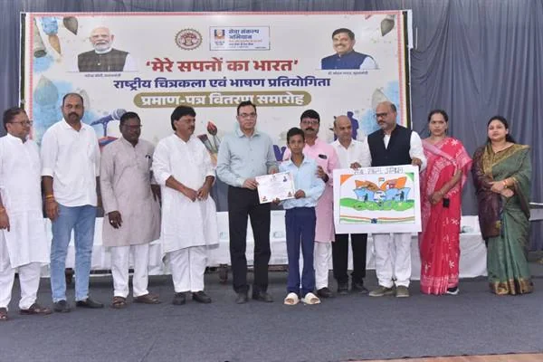मंच पर बैनर के सामने खड़े जनप्रतिनिधि और अधिकारी एक विद्यार्थी को प्रमाण पत्र दे रहे हैं, पास में एक व्यक्ति बच्चों द्वारा बनाया रंगीन चित्र दिखा रहा है।